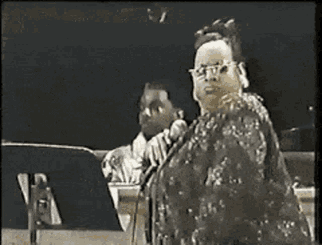 a woman singing into a microphone with a man playing a piano in the background .