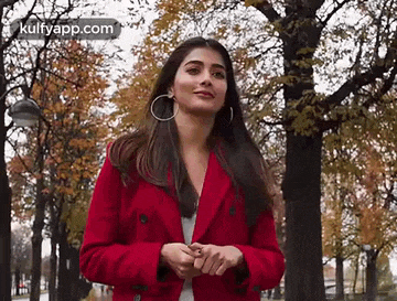 a woman wearing a red coat and earrings is standing in a park .