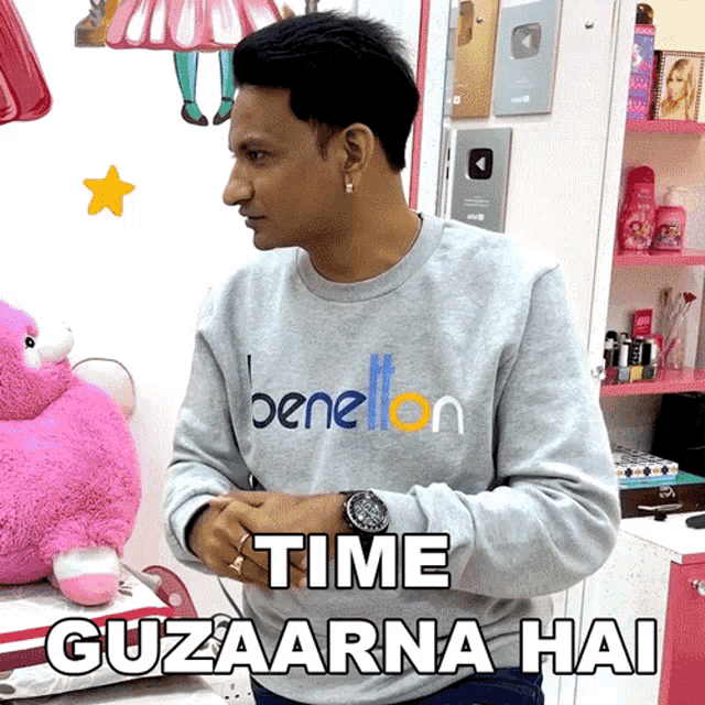 a man wearing a grey benetton sweatshirt stands in front of a pink stuffed animal