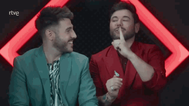 two men are laughing in front of a red background and the word rtve is on the bottom