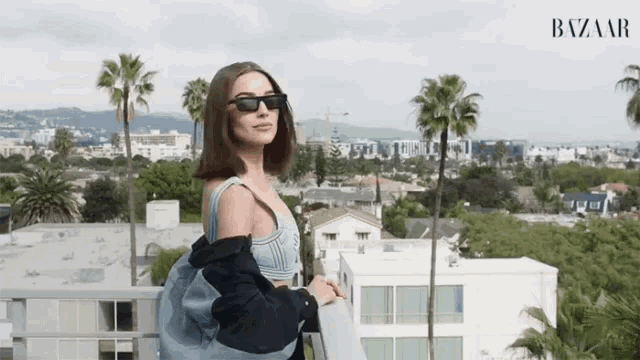 a woman wearing sunglasses is standing on a balcony overlooking a city with the word bazaar on the bottom right