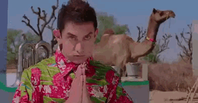 a man in a floral shirt is praying in front of a camel in the desert .