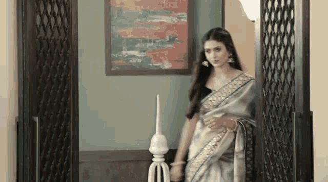 a woman in a saree is standing in a room with a picture on the wall .