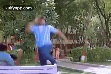 a man in a blue shirt is standing on a bench in a park with his hands in the air .
