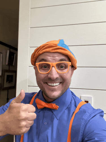 a man in a blue shirt and orange suspenders is wearing a hat and glasses and giving a thumbs up