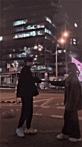 a man and a woman are walking down a street at night with a building in the background that says " you "