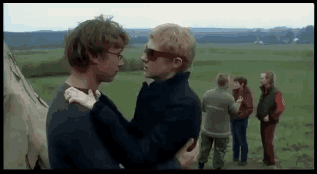a group of people are standing in a field talking to each other . one of the men is wearing sunglasses .
