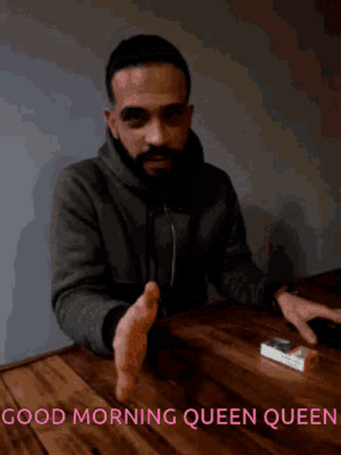 a man with a beard is sitting at a table with a cigarette in his hand and the words good morning queen queen below him