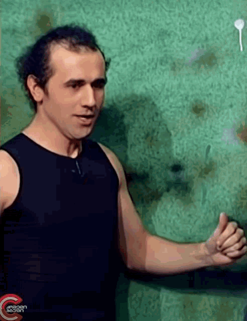 a man in a black tank top is giving a thumbs up in front of a green wall