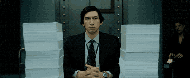 a man in a suit and tie is sitting in front of stacks of paper