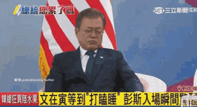 a man in a suit and tie is sitting in front of a flag