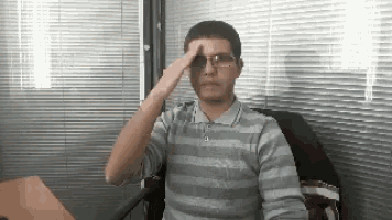 a man wearing glasses and a striped shirt salutes while sitting at a desk .