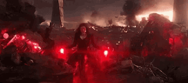 scarlet witch is holding two red lights in her hands while standing in front of a destroyed building .