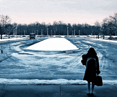 a woman standing on a sidewalk in front of a snow covered parking lot