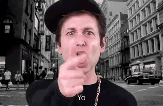 a man wearing a hat and a black shirt is pointing at the camera with the word yo in front of him .