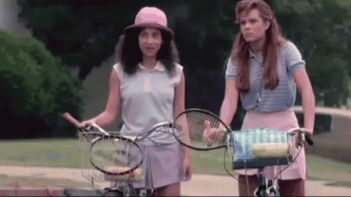 two women are standing next to each other on a sidewalk holding tennis racquets .