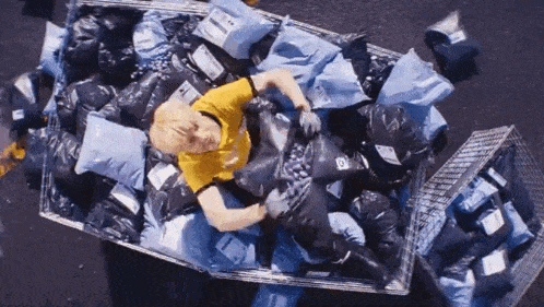 a man in a yellow shirt is laying in a coffin full of trash bags