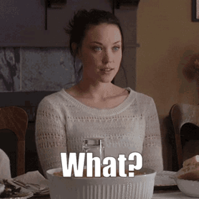 a woman in a white sweater is sitting at a table with a bowl of food and a sign that says what ?