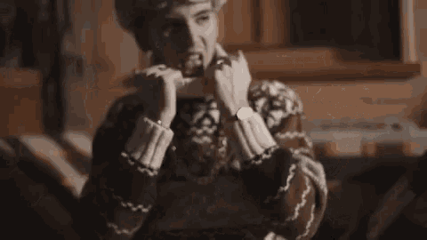 a man wearing a sweater and a fur hat is sitting on a couch and covering his face with his hands .