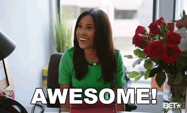 a woman in a green shirt is sitting at a desk with a vase of red roses in front of her and the words awesome on the screen