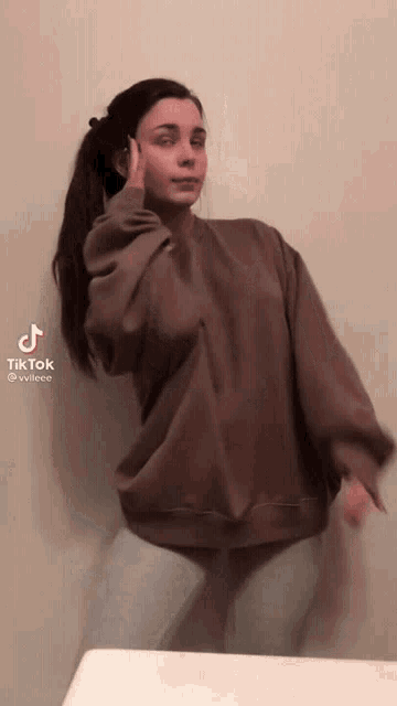 a woman in a brown sweater is standing in front of a wall with a tiktok sticker on her face