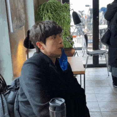 a man in a black coat sits at a table with a blue bag in his mouth