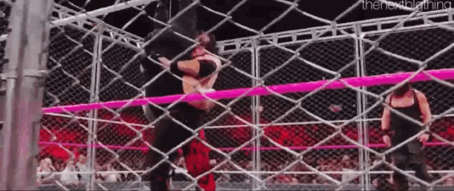 a wrestler is jumping over a chain link fence while another wrestler watches .
