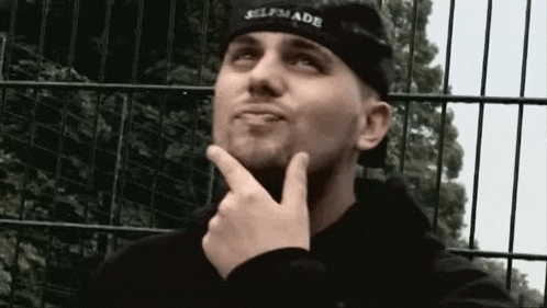 a man wearing a black hat and a black shirt is standing in front of a fence with his hand on his chin .