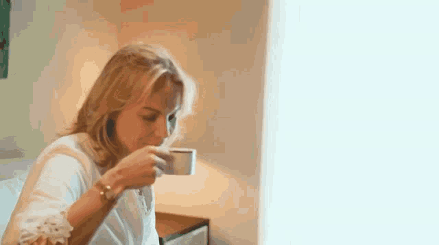a woman is drinking a cup of coffee while sitting at a table in front of a window .
