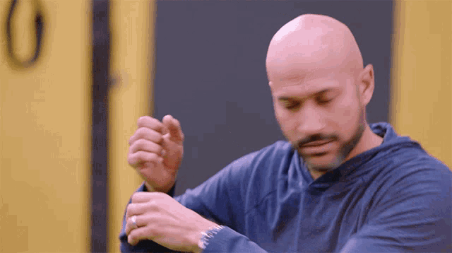 a bald man with a beard wearing a blue hoodie looks at his watch