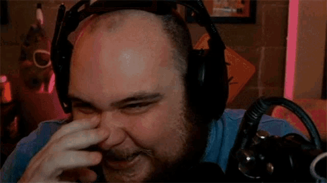 a bald man with a beard wearing headphones is covering his nose while laughing in front of a microphone .