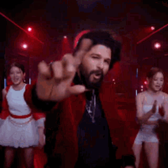 a man in a red jacket is pointing at the camera while two women in white dresses are dancing behind him .