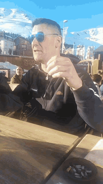 a man wearing sunglasses sits at a table with a bowl of cigarettes on it