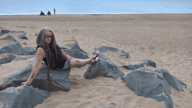 a woman with dreadlocks sits on some rocks on the beach