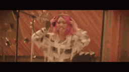 a woman with pink hair and headphones is singing into a microphone in a recording studio .