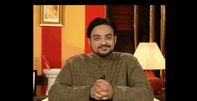 a man with glasses and a beard is sitting with his hands folded in front of a lamp