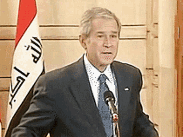 a man in a suit and tie stands in front of a microphone in front of a flag with arabic writing