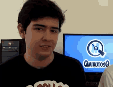 a man wearing a shirt that says qminutosq stands in front of a computer screen