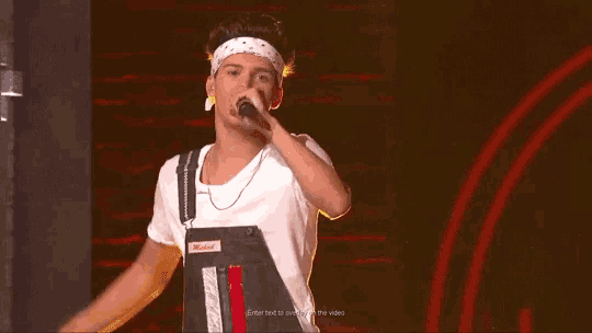 a young man wearing overalls and a headband is singing into a microphone on a stage .