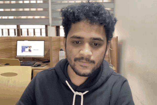 a man wearing a black hoodie looks at the camera with a computer monitor in the background