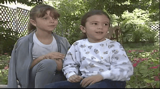 two young girls are sitting next to each other in front of a garden