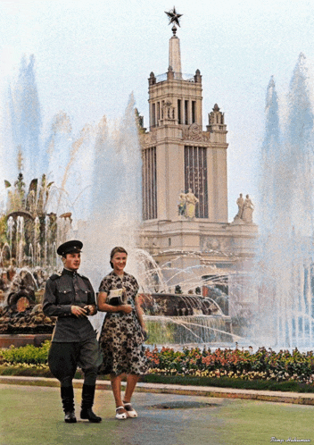 a man and a woman standing in front of a fountain with a building in the background