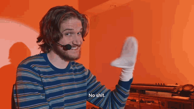 a man wearing a microphone and a striped sweater holds up a sock and says no shit