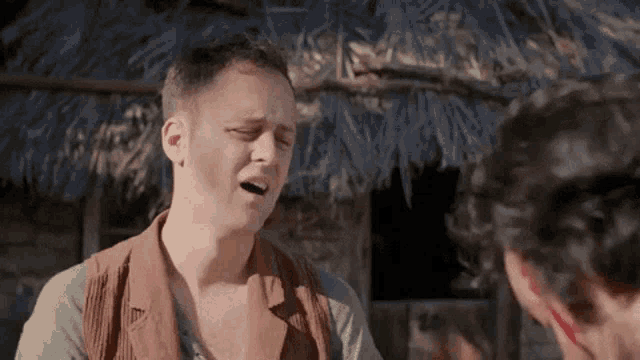 a man is making a funny face while talking to another man in front of a thatched hut .