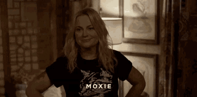 a woman wearing a moxie t-shirt is standing in a room with her hands on her hips and smiling .