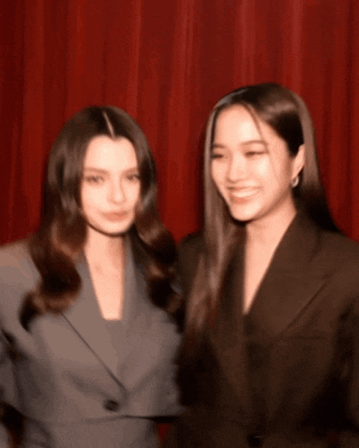 two women standing next to each other with a red curtain behind them