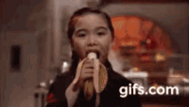 a young girl is eating a banana in a kitchen in a gif .