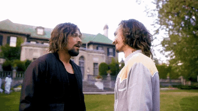 two men are standing next to each other in front of a large house and talking to each other .