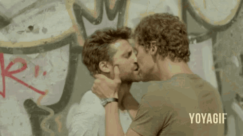 two men are kissing in front of a wall with graffiti on it and the words yoyagii on the bottom