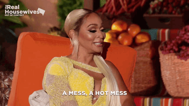 a woman in a yellow dress says " a mess a hot mess " while sitting in an orange chair